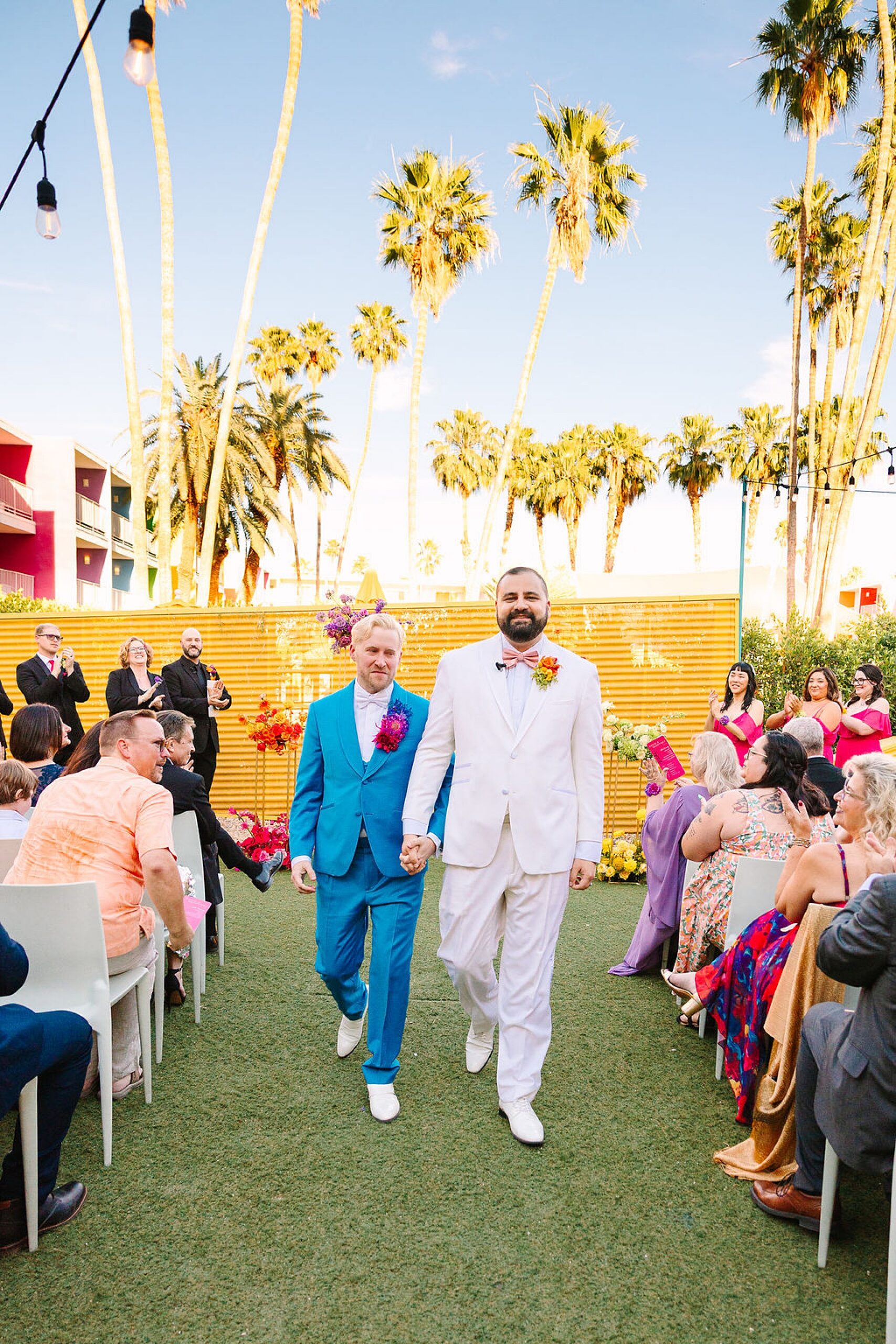 Grooms cry while walking up the aisle holding hands under palm trees during their the saguaro palm springs wedding