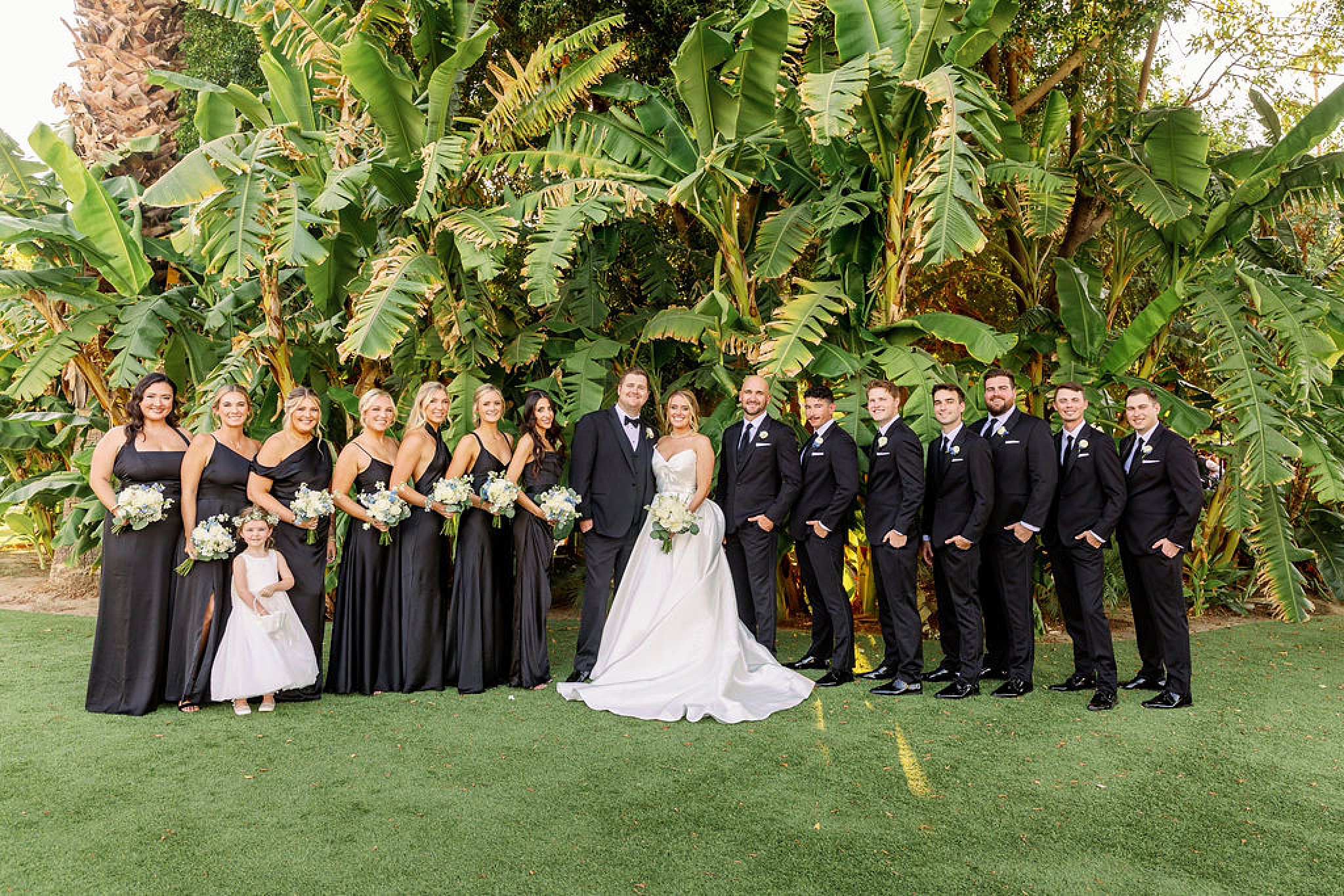 Happy newlyweds stand smiling with their wedding party in all black in a tropical garden