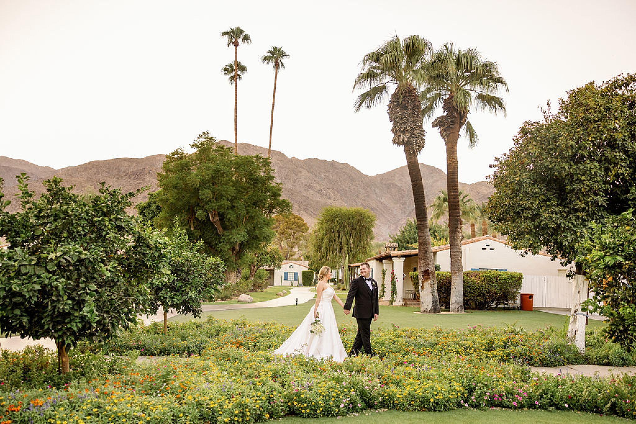 A bride and groom walk holding hands through a flower garden path at sunset after working with weddings with pam olson