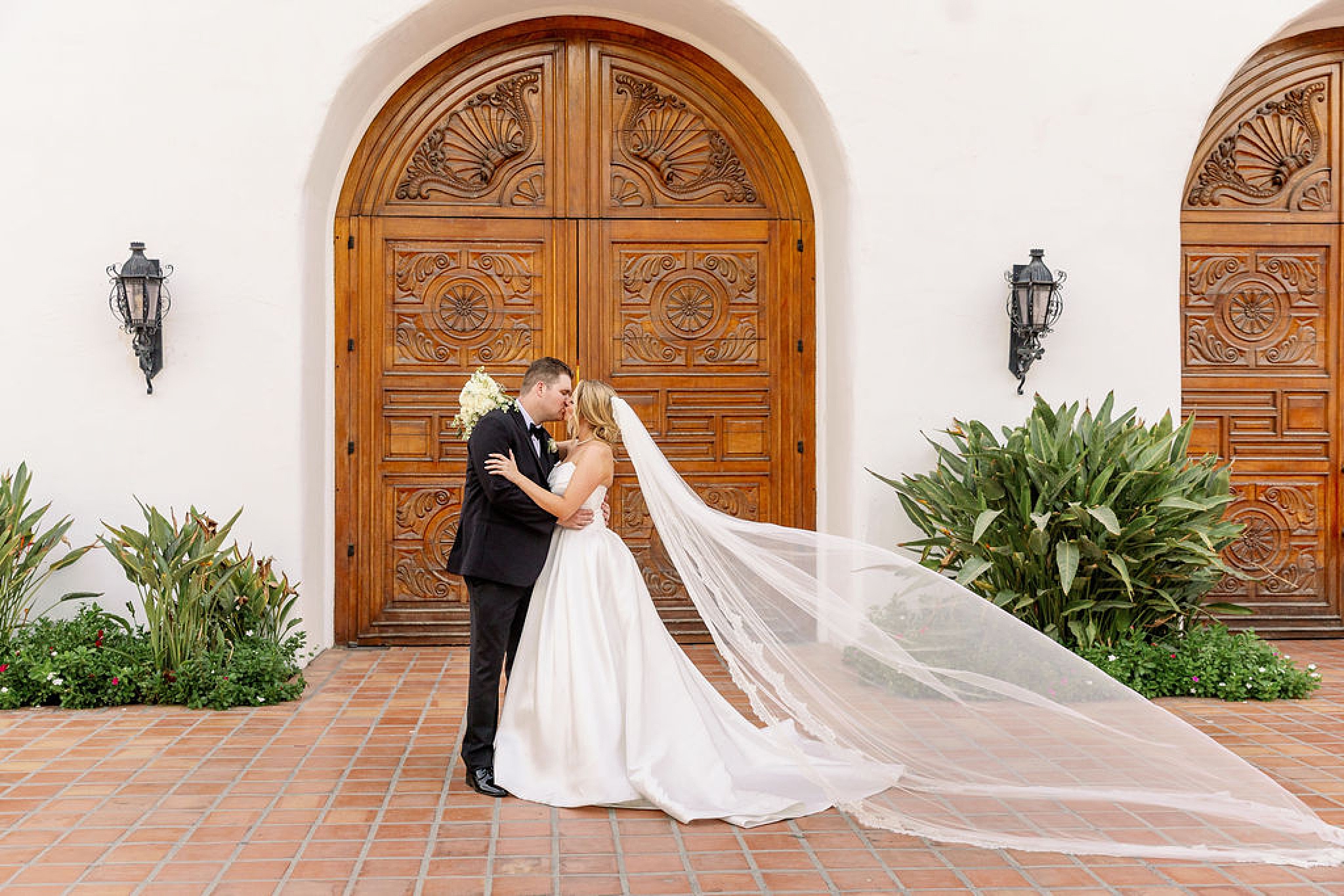 A bride and her groom in black tux kiss with the long veil flowing in front of large ornate wooden doors while enjoying their day planned by weddings with pam olson