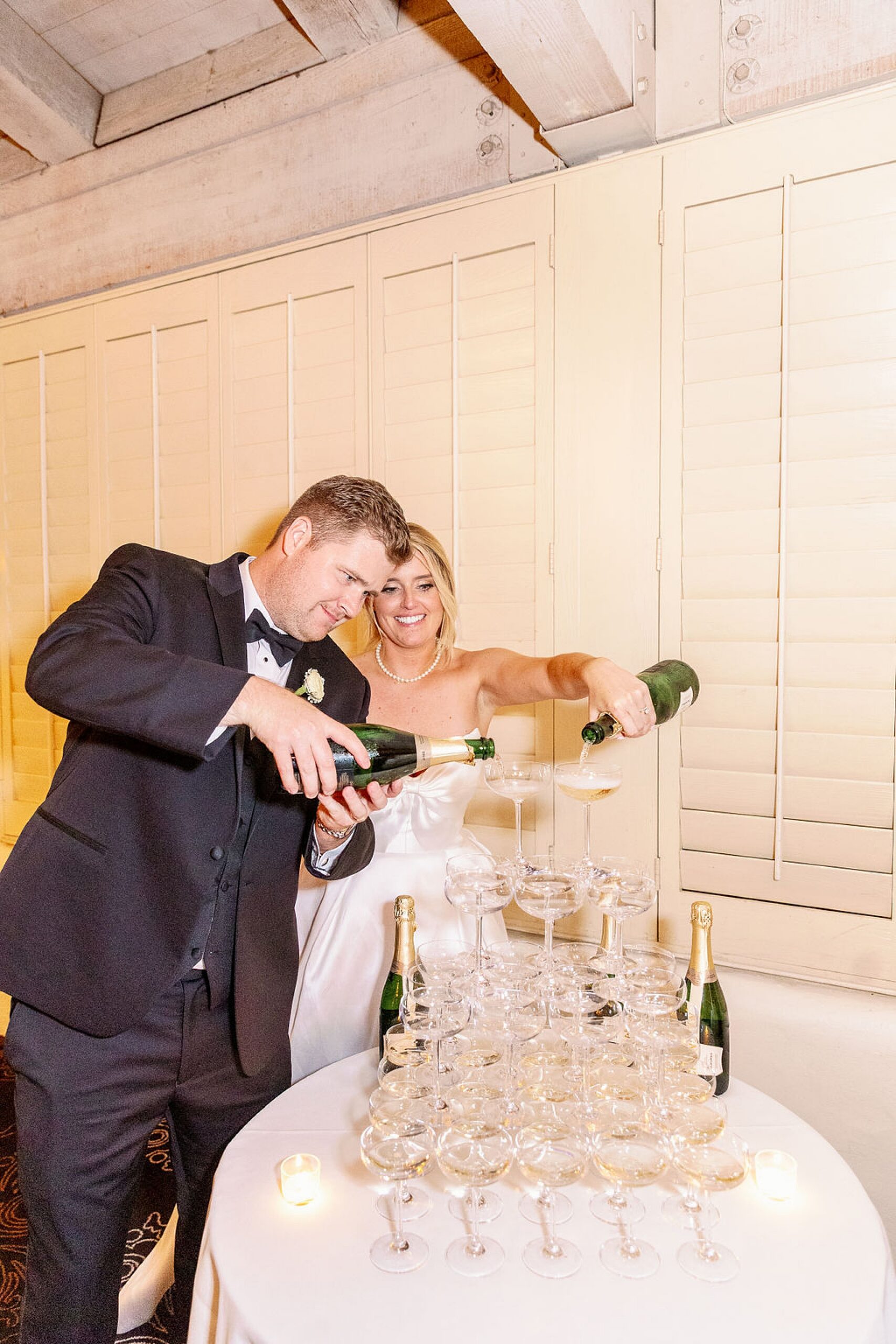 Happy newlyweds pour champagne over a tower of glasses on a white table